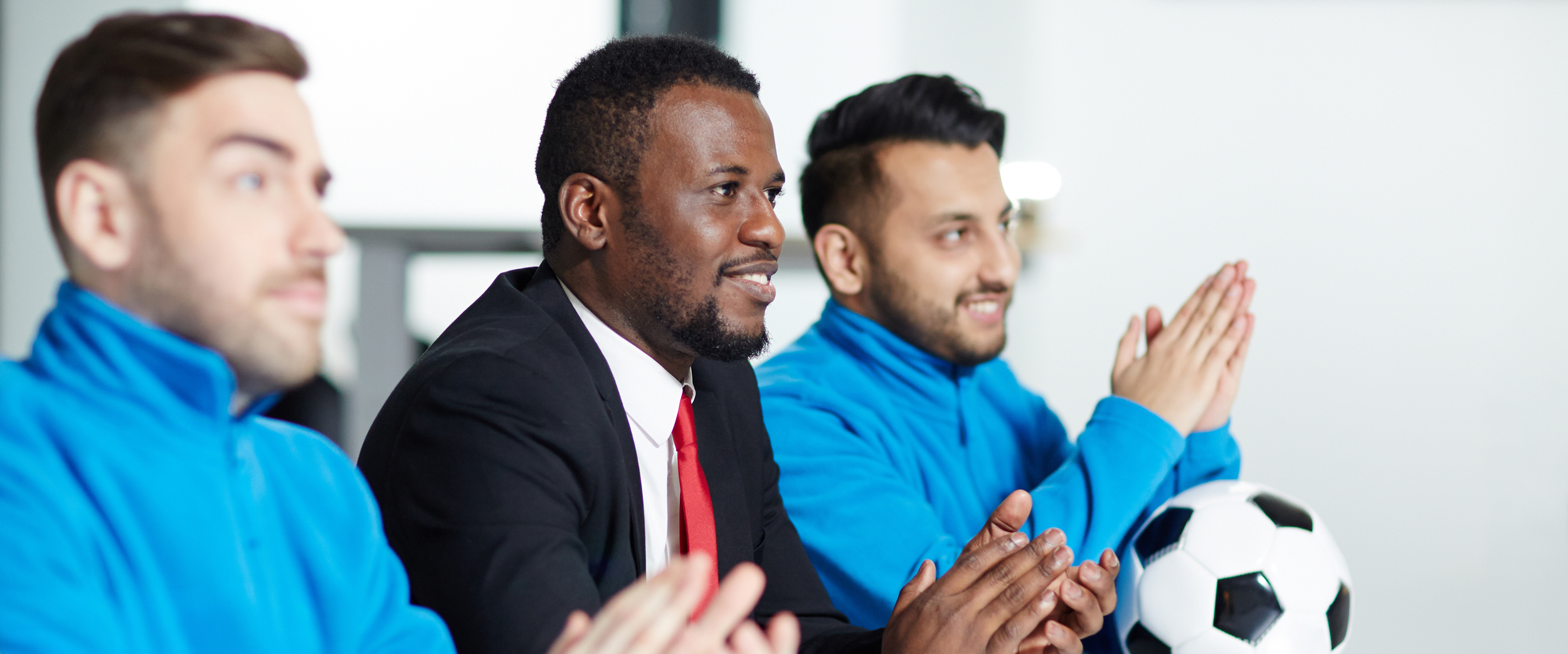 Three individuals sitting at a conference table, two in athletic warm-ups, one in a business suit, with a soccer ball on the table.
